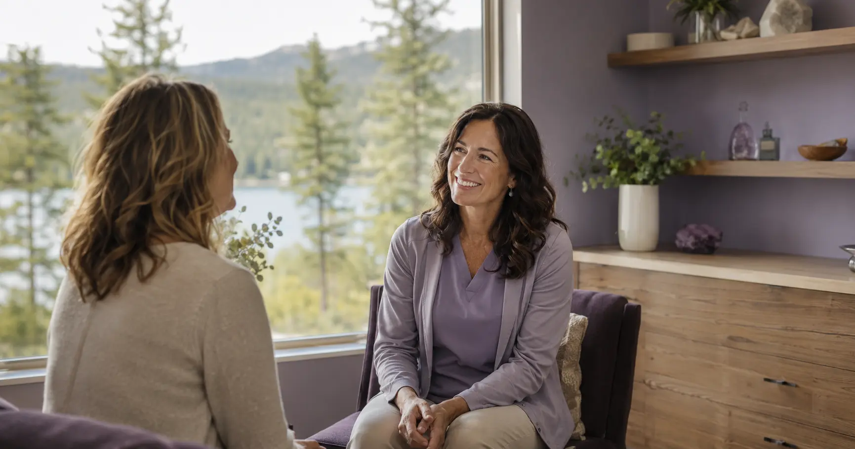 A warm, credentialed female clinician welcomes a woman in her fifties into a bright mountain wellness clinic in Evergreen, CO, with tall evergreen pines and a glimpse of an alpine lake through the window, conveying honest, local longevity and IV therapy care.